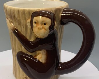 May include: A ceramic coffee mug with a monkey design. The mug features a brown monkey clinging to a tree trunk-shaped mug. The handle is a curved, dark brown shape. The mug has a light beige exterior and a white interior.