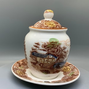May include: A white ceramic jar with a lid and matching saucer. The jar and lid feature a brown and yellow floral pattern. The jar has a landscape scene with a house, trees, and a green sun. The saucer has a matching floral pattern.