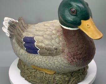 May include: A ceramic duck figurine with a green head, brown body, and gray wings. The duck has a yellow beak and is resting on a nest made of faux grass. The duck's wings have blue and white accents.