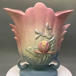 May include: A ceramic vase featuring a scalloped rim and a gradient color scheme transitioning from pink at the top to cream in the center and blue at the base. The front displays a raised relief design of a pink flower with green leaves.
