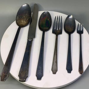 May include: A set of six tarnished silver-toned utensils including a large spoon, a dinner knife, a medium spoon, a four-tined fork, a small spoon, and a tiny two-tined fork, all featuring a tapered handle design with a pointed end.