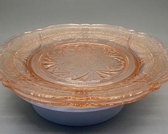 May include: A pink glass plate with a delicate floral pattern. The plate has a scalloped edge and is made of pressed glass.