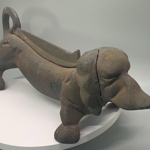 May include: A cast iron dachshund dog doorstop in a weathered brown finish. The dog is depicted in a prone position with a long body, short legs, and a curved tail. The doorstop measures approximately 12 inches in length.