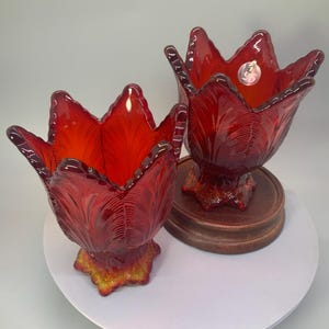 May include: Two ruby red glass tulip-shaped candle holders. The candle holders have a detailed leaf pattern and sit on a wooden base. The glass has a glossy finish and a vibrant color.