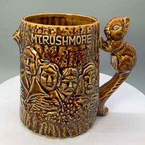 May include: Brown ceramic mug with a tree trunk design, featuring the faces of Mount Rushmore. The handle is a sculpted squirrel. The mug is inscribed with "MT. RUSHMORE".