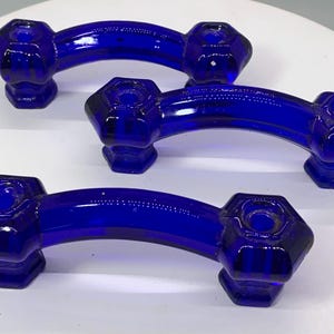 May include: Three cobalt blue glass cabinet pulls. Each pull has a curved handle and hexagonal end pieces. The glass is transparent, allowing light to pass through. These vintage-style pulls would be suitable for a kitchen or bathroom.