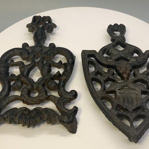 May include: Two antique cast iron trivets. One is a shield shape with an emblem, the other is an openwork design. Both are dark brown with intricate patterns and details, perfect for kitchen decor.