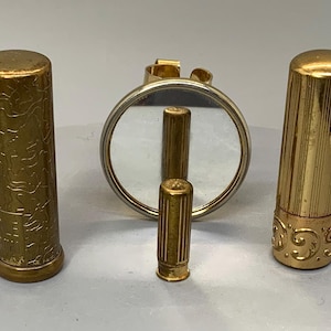 Puede incluir: Tres envases de lápiz labial vintage de color dorado con un pequeño espejo redondo. Los envases de lápiz labial tienen diseños diferentes y están todos cerrados.