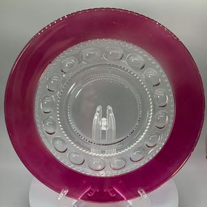 May include: A clear glass plate with a raised, textured rim and a pink rim around the edge. The plate has a pattern of small circles around the center.