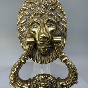 May include: A brass lion-head door knocker. The knocker features an oval backplate with a detailed lion's face, a handle, and a decorative lower section with a smaller face. The metal has a polished, antique finish.