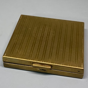 May include: A gold-colored metal compact with a ridged surface. The compact is closed and has a small latch on the side.