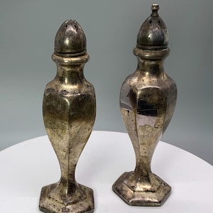 May include: Two antique silver-colored salt and pepper shakers. Each shaker has a bulbous body, a hexagonal base, and a conical top with small holes. The shakers have a tarnished appearance, suggesting age and use. They are displayed on a white surface.
