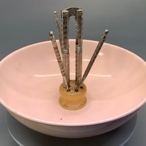 May include: A vintage nutcracker set with a silver-toned nutcracker and picks, displayed in a light pink serving bowl. The nutcracker and picks are held in a small, light-colored holder. The bowl is round and has a glossy finish.