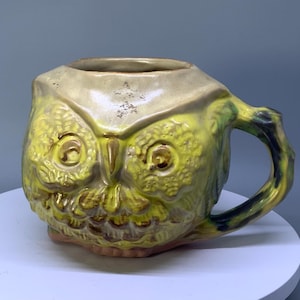 May include: A ceramic mug shaped like an owl with a glossy yellow and green glaze. The owl features textured feathers, large round eyes, and a pointed beak. A curved handle extends from the side, and the top rim has a smooth, light beige finish.