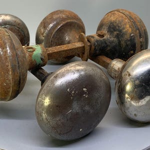 May include: A collection of vintage door knobs in various states of wear. The knobs are primarily metallic, with shades of brown, silver, and rust. Some knobs show signs of age with rust and discoloration, while others retain a polished appearance.