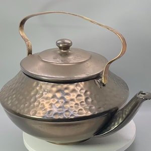 May include: A metallic, dark-toned teapot featuring a hammered, dimpled texture across its rounded body. It includes a matching lid with a central knob, a curved spout, and a thin, arched handle that extends upward from the sides of the vessel.