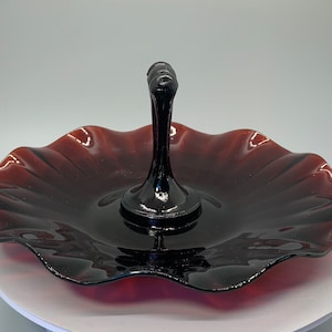 May include: A dark red glass serving dish with a scalloped edge and a black handle. The dish is perfect for serving snacks or desserts.