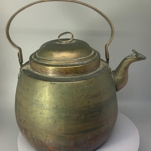 May include: A large, antique brass kettle with a rounded body, a hinged lid, and a curved handle. The kettle has a spout shaped like a bird's head.