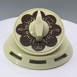 May include: A vintage kitchen timer in cream and brown. The timer features a circular face with a floral design and numbers indicating minutes. The dial reads "MARK TIME" and has a cream-colored knob. The base has a series of rectangular cutouts.