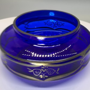 May include: A cobalt blue glass bowl with gold trim and floral designs. The bowl has a wide, rounded base and a narrower opening. The gold trim outlines the rim and base, with decorative floral patterns. The bowl is likely vintage.