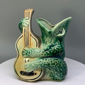 May include: A ceramic vase shaped like a green, speckled frog playing a light tan acoustic guitar. The frog has its mouth open, and its hands are positioned on the neck and body of the instrument. The piece features a glossy finish.