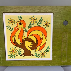May include: A green wooden frame with a white tile featuring a colorful rooster with a yellow, orange, and brown design. The rooster is surrounded by yellow flowers and green leaves. The frame has a small, round, black knob on the right side.
