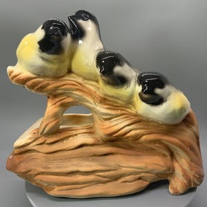 May include: A ceramic sculpture of three birds perched on a stylized tree branch. The birds have black and white heads with yellow accents. The branch is brown with a textured, wood-like appearance. The sculpture is likely a decorative item.