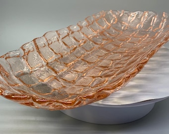 May include: A clear, rectangular glass serving dish with a textured, grid-like pattern and a ruffled edge. The dish has a light peach hue and is displayed on a white surface. This decorative piece is suitable for serving food.