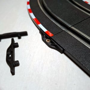 Könnte beinhalten: Ein Abschnitt einer schwarzen Autorennbahn mit weiß-roten Rennstreifen. Mehrere schwarze Kunststoff-Streckenverbinder sind in der Nähe verstreut. Die Strecke hat zwei parallele silberne Rillen.