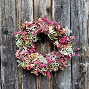 Könnte beinhalten: Ein kreisförmiger Kranz aus leuchtend rosa Heidekraut, getrockneten burgunderroten Hortensienblüten, kleinen weißen Schleierkrautblüten und silbrig-grünen Flechtenzweigen. Das Blumenarrangement ist dicht gefüllt und wirkt sehr strukturiert.