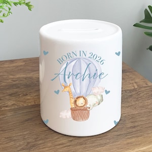 May include: White ceramic money bank with a slot on top. The bank features a blue hot air balloon design with a giraffe, lion, and elephant in a basket. Text reads "BORN IN 2026 Archie" with blue hearts.