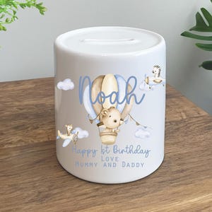 May include: White ceramic money box with a blue and brown hot air balloon design. The balloon has a small animal inside and is surrounded by clouds. The text "Noah" is written in blue on the balloon. The text "Happy 1st Birthday Love Mummy and Daddy" is written in blue at the bottom of the box.