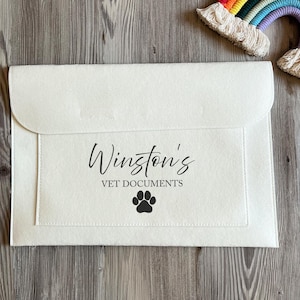 May include: Cream-coloured felt document holder with a flap closure. The front displays the text "Winston's VET DOCUMENTS" in black script, and a black paw print. The holder is designed for pet documents. A rainbow decoration is in the background.