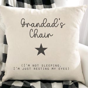 May include: Cream-coloured square cushion with the text "Grandad's Chair" in a dark grey script font, a black star, and the phrase "(I'M NOT SLEEPING, I'M JUST RESTING MY EYES)" in a sans-serif font. The cushion is on a white surface.
