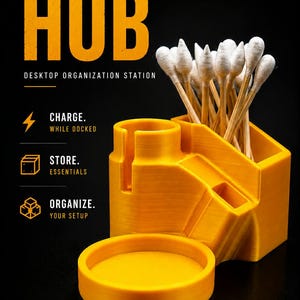 May include: A bright yellow desktop organization station labeled PIVOT HUB with a circular tray, a vertical slot, and a rectangular compartment holding several cotton swabs. The text reads PIVOT HUB DESKTOP ORGANIZATION STATION, CHARGE WHILE DOCKED, STORE ESSENTIALS, ORGANIZE YOUR SETUP, and DF BUILT DIFFERENT.