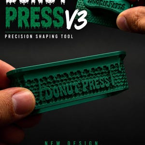 May include: A dark green donut press tool featuring a textured honeycomb pattern on the exterior walls with the words DONUT PRESS embossed on the side. The tool has a matching green handle with the text DOOBIE FATES printed on the top surface.