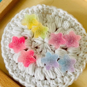 May include: A white crocheted coaster with colourful star-shaped sweets on top. The sweets are in shades of pink, yellow, blue, and purple.