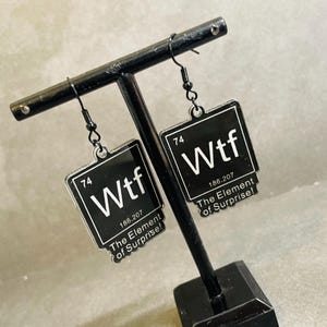 May include: A pair of black dangle earrings with a square pendant featuring the word "Wtf" and the text "The Element of Surprise!"  The pendant is styled to look like a periodic table element with the number "74" and the atomic weight "186.207".