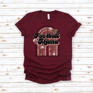 May include: Burgundy t-shirt with a retro-style graphic that says "Football Mama" in black text. The graphic features a football, a helmet, flowers, and peace signs.