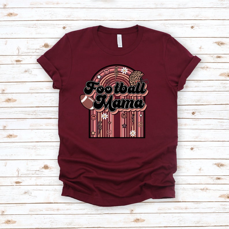May include: Burgundy t-shirt with a retro-style graphic that says "Football Mama" in black text. The graphic features a football, a helmet, flowers, and peace signs.