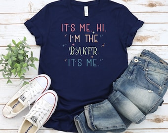 Peut inclure: Un t-shirt bleu marine avec le texte "It's me. Hi. I'm the baker. It's me." en rose, bleu et blanc. Le t-shirt est posé sur une surface en bois blanc avec une paire de baskets blanches et un jean bleu.