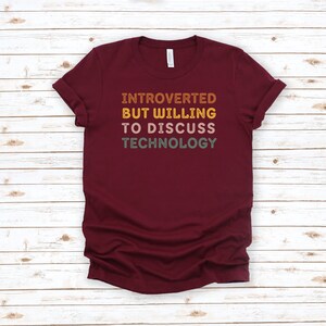 May include: A maroon t-shirt with the text "Introverted But Willing To Discuss Technology" in a retro font with colorful accents.