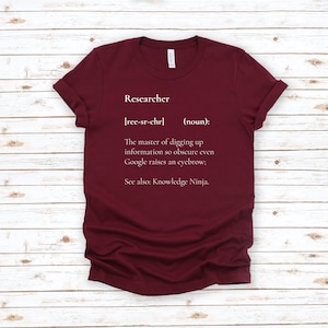 May include: A maroon t-shirt with white text that reads "Researcher [ree-sr-chr] (noun): The master of digging up information so obscure even Google raises an eyebrow; See also: Knowledge Ninja."