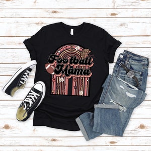 May include: Black t-shirt with a pink and brown graphic design featuring the text "Football Mama" in a retro font. The graphic includes a football, daisies, peace signs, and a rainbow.