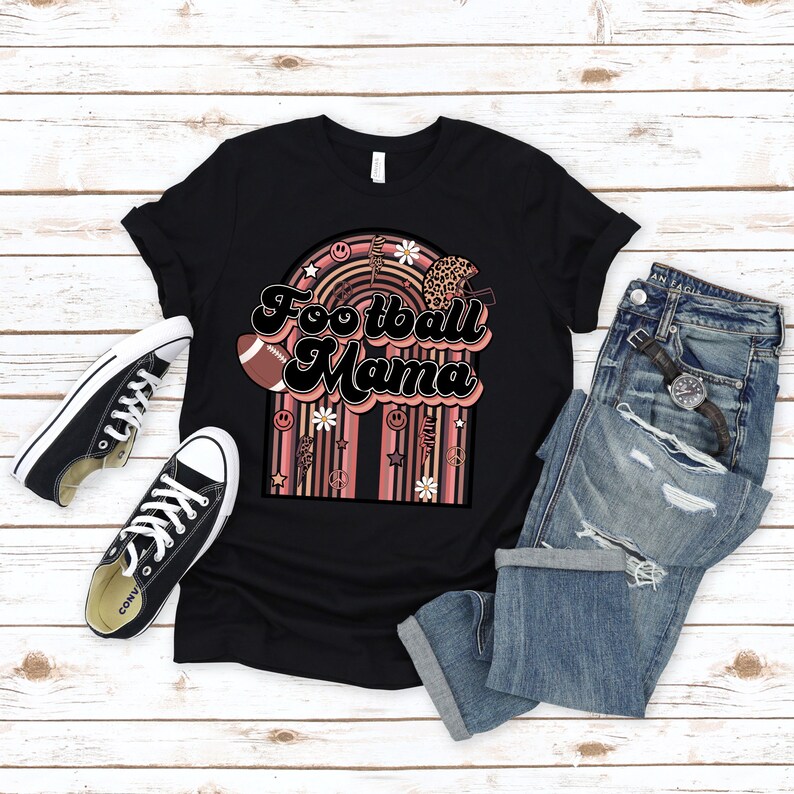 May include: Black t-shirt with a pink and brown graphic design featuring the text "Football Mama" in a retro font. The graphic includes a football, daisies, peace signs, and a rainbow.