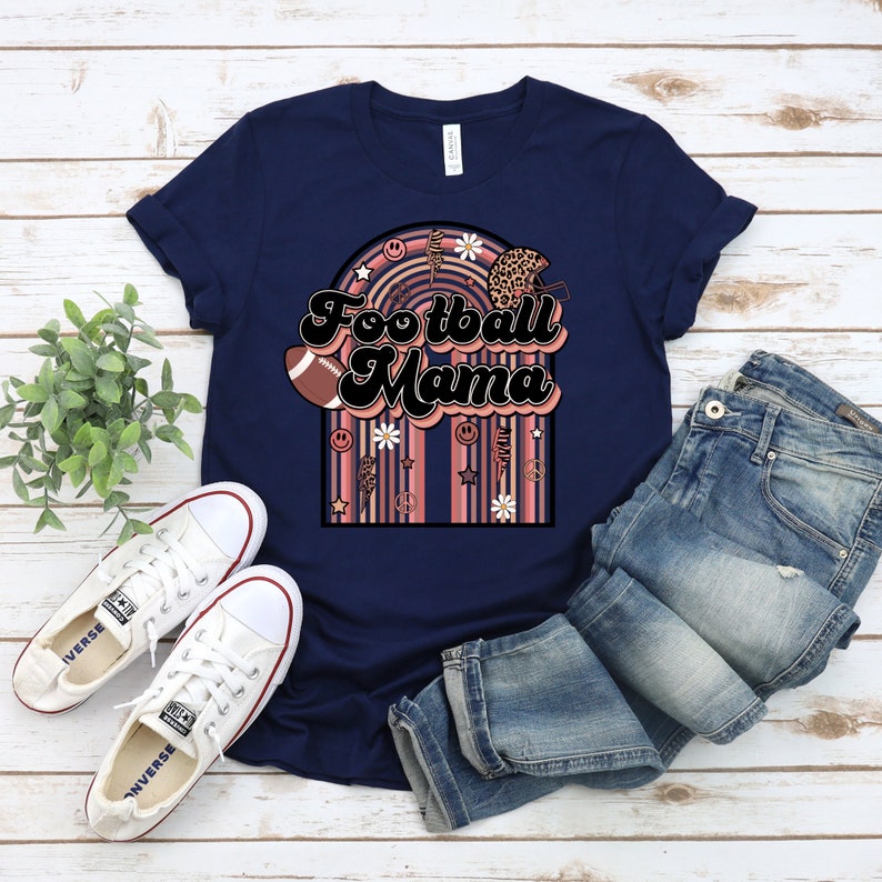May include: Navy blue t-shirt with a retro-style graphic that says "Football Mama" with a football, daisies, and peace signs.