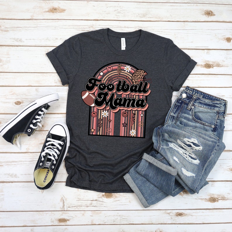 May include: A dark grey t-shirt featuring the text "Football Mama" in a retro font. The text is surrounded by a rainbow arch with various patterns, including flowers, peace signs, and stars.