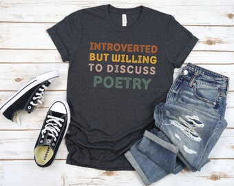 Op de afbeelding: Een donkergrijs t-shirt met de tekst "Introvert maar bereid om over poëzie te praten" in kleurrijke letters. Het shirt is gestyled met een distressed denim jeans en zwarte sneakers.