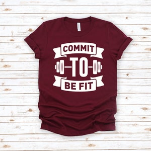 Op de afbeelding: Een maroon t-shirt met een witte afbeelding met de tekst "Commit to Be Fit" met een halterstangontwerp.