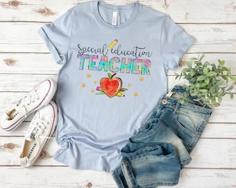 Può includere: Una maglietta blu chiaro con il testo "Special Education Teacher" in colori arcobaleno con una mela rossa e matite. La maglietta è su una superficie di legno con un paio di scarpe da ginnastica bianche e jeans blu.